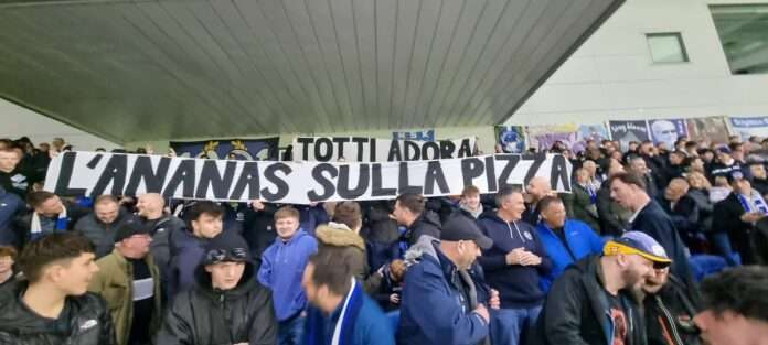 Brighton fans respond to Roma's distasteful queen-related message with a clever banner, humorously associating Totti with pineapple pizza, blending royal tribute with football banter